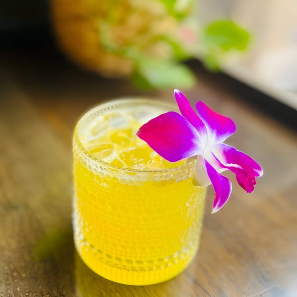 Vibrant yellow Thai cocktail with ice, garnished with a bright purple orchid, on a wooden table.