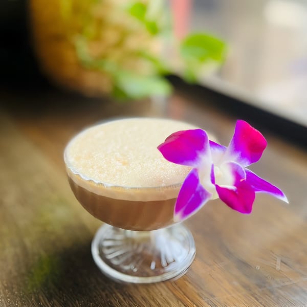 A Thai iced tea cocktail with a creamy top, garnished with a vibrant purple orchid.