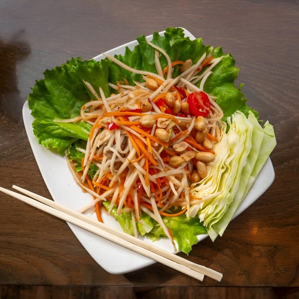 Som Tum (papaya salad) with peanuts, tomatoes, and carrots, served with lettuce and chopsticks.